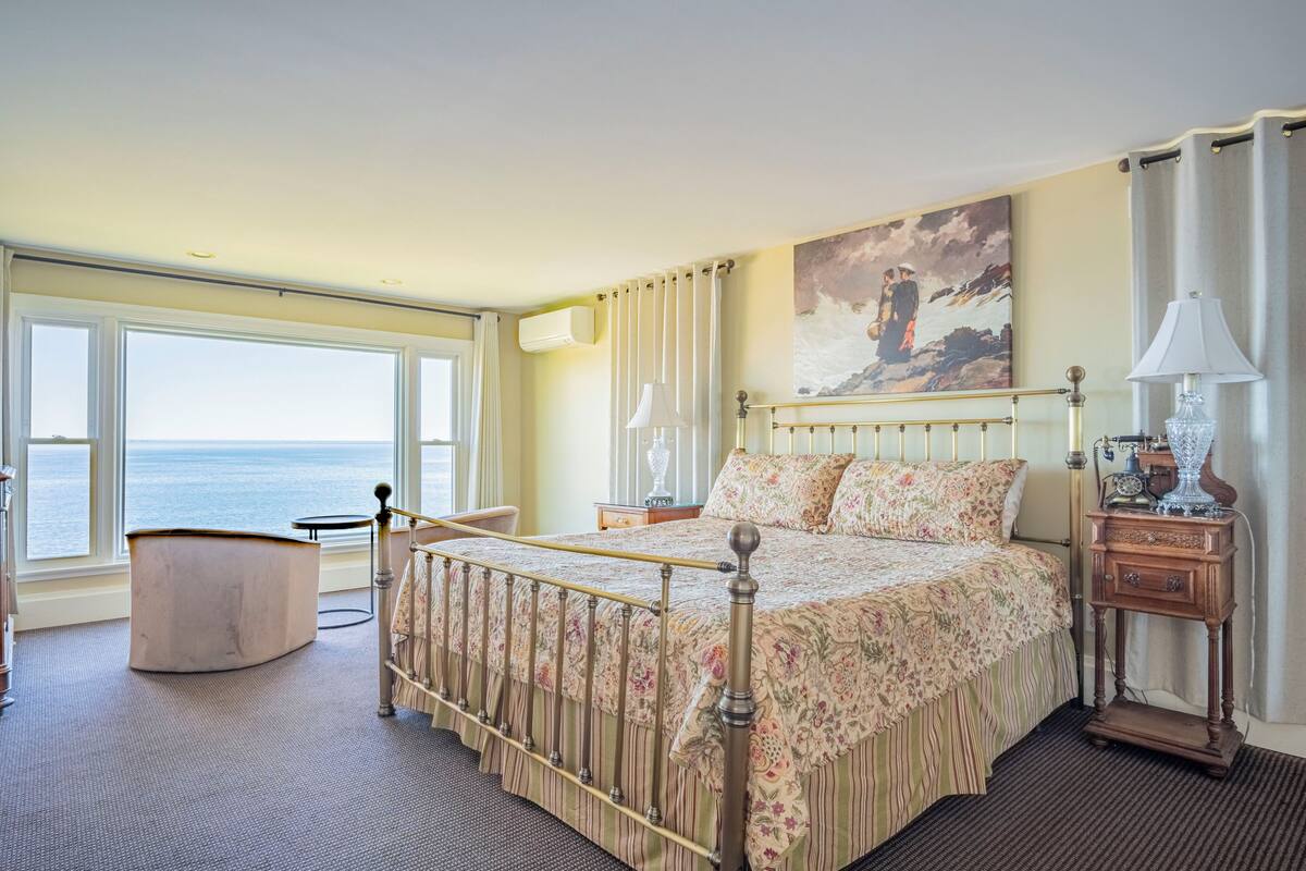 Un dormitorio con cama, mesita de noche, lámpara, un cuadro y vistas al mar.