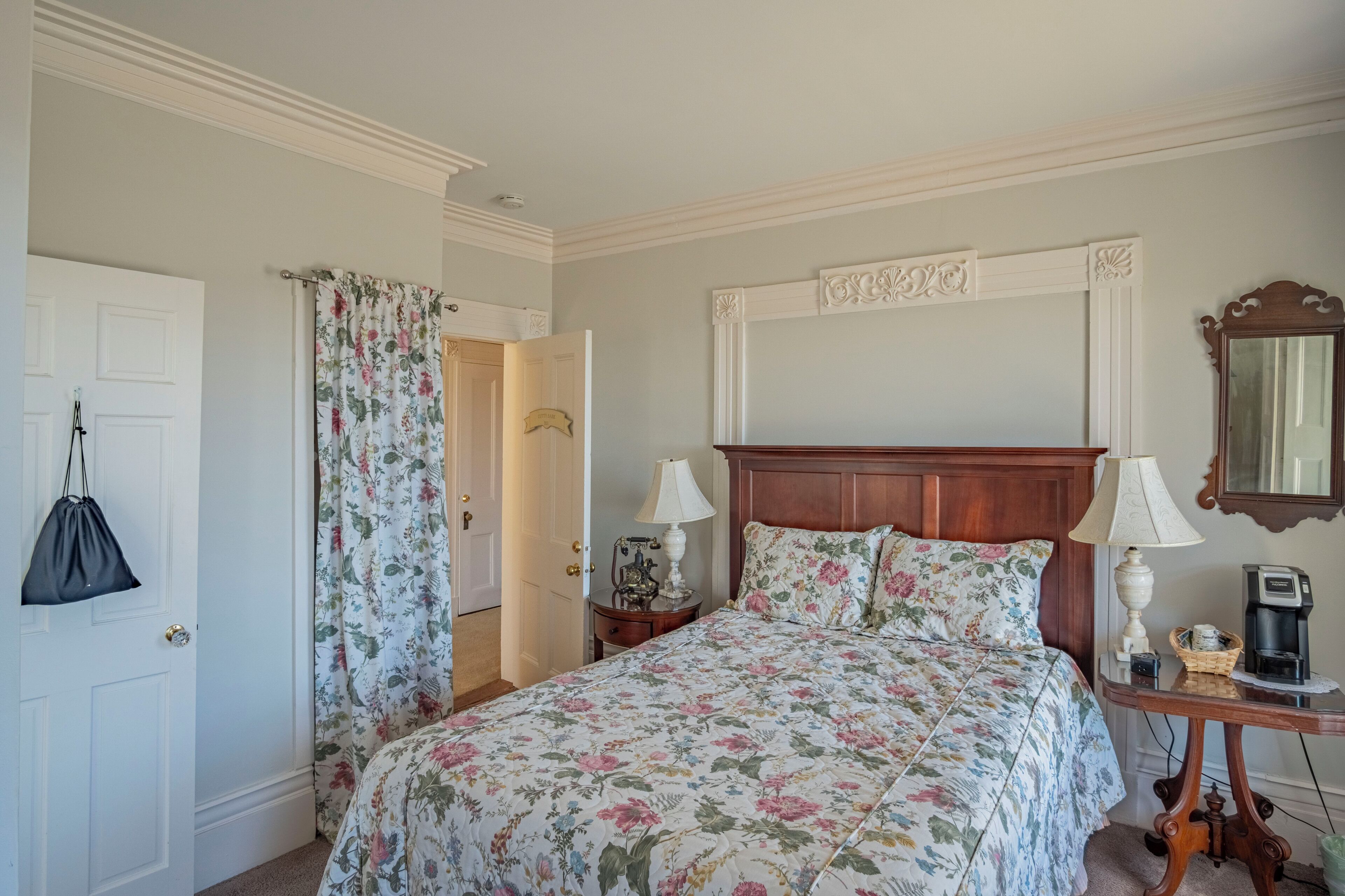 Een slaapkamer met een bloemenprint bedspreider, een houten hoofdbord, twee bedlampen, een nachtkastje met een lamp, een koffiezetapparaat en een spiegel aan de muur.