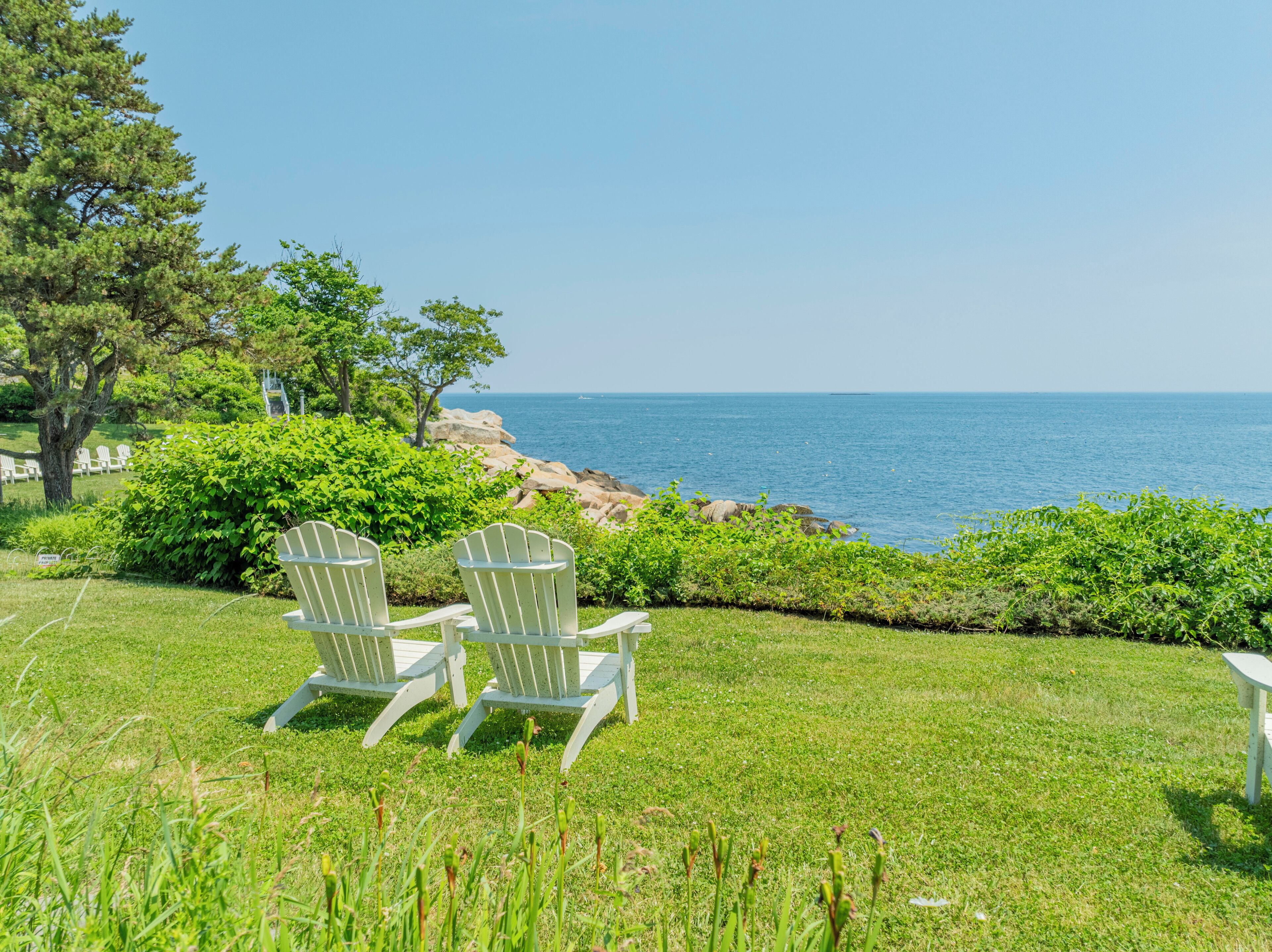 A grassy area with two Adirondack chairs facing a rocky coastline and the sea.