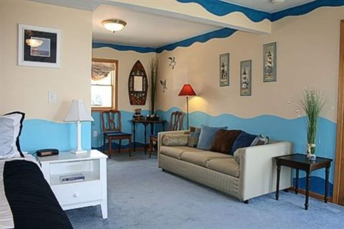 A cozy living room with a sofa, a small table, and a lamp. There are framed pictures on the wall and a window with curtains.