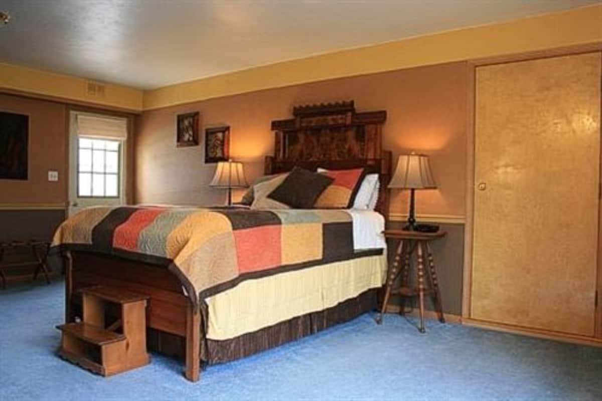 A bedroom with a large bed, two bedside lamps, a wooden headboard, a wardrobe, and a window with blinds.