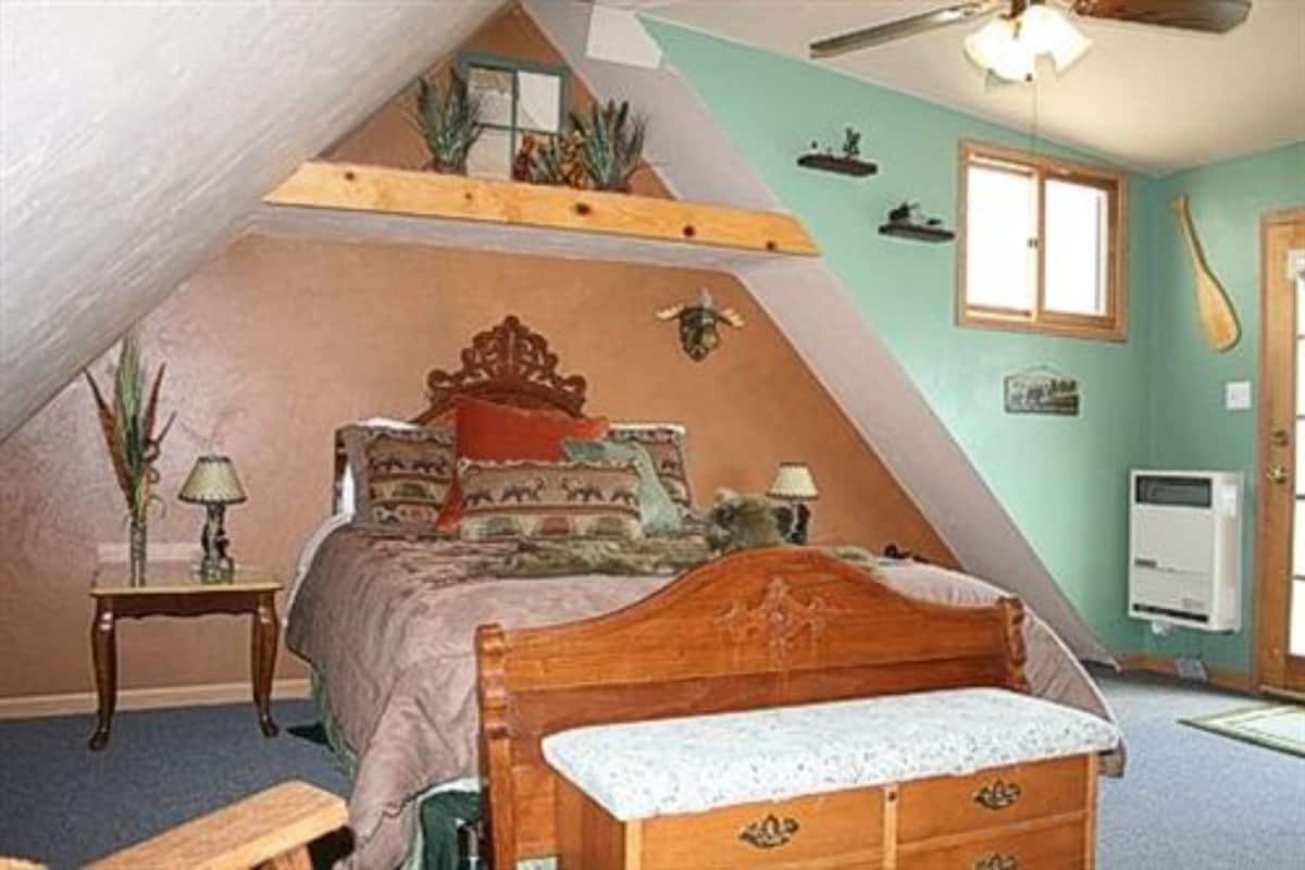 A bedroom with a sloped ceiling, a wooden bed, a small bedside table, a lamp, and a wall-mounted heater.