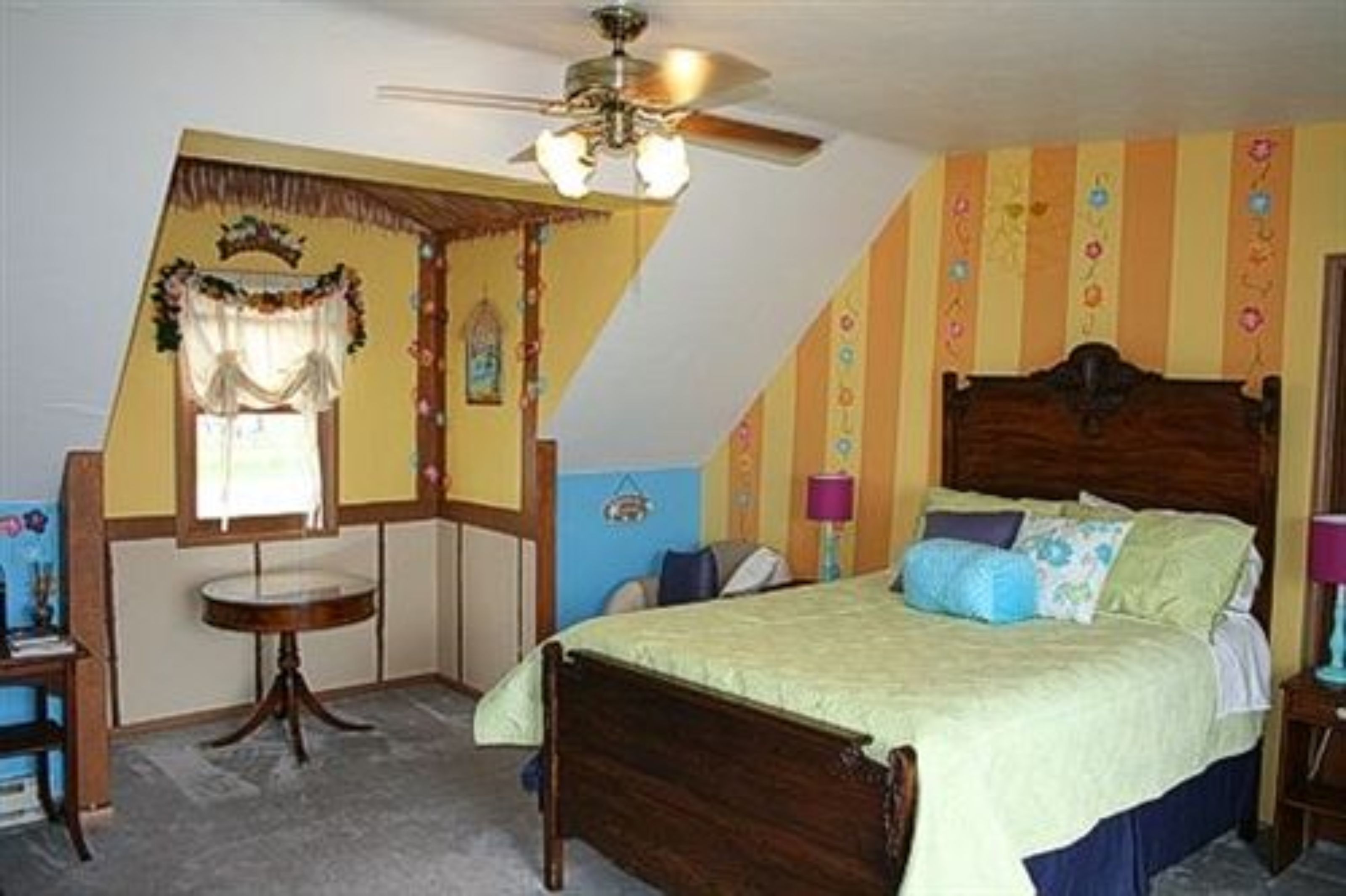 A bedroom with a bed, a ceiling fan, a window with curtains, and a small table.
