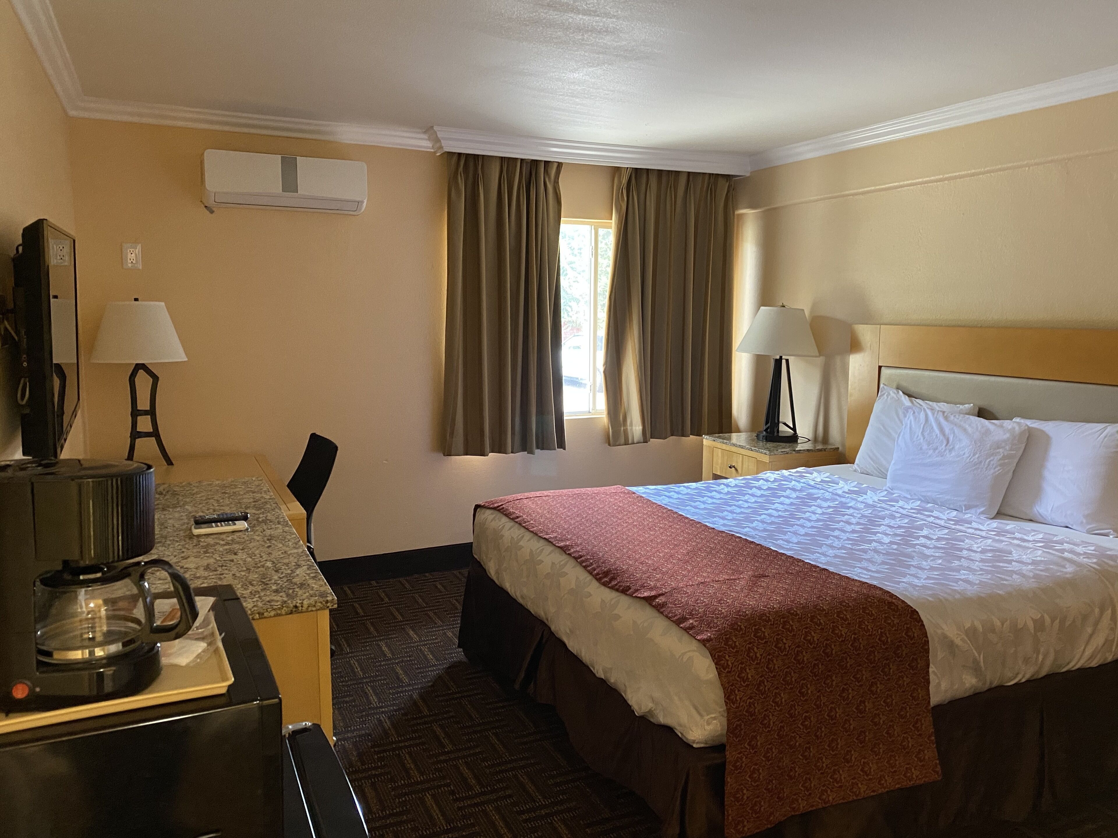 A hotel room with a bed, a desk with a coffee maker, a TV, and a window with curtains.