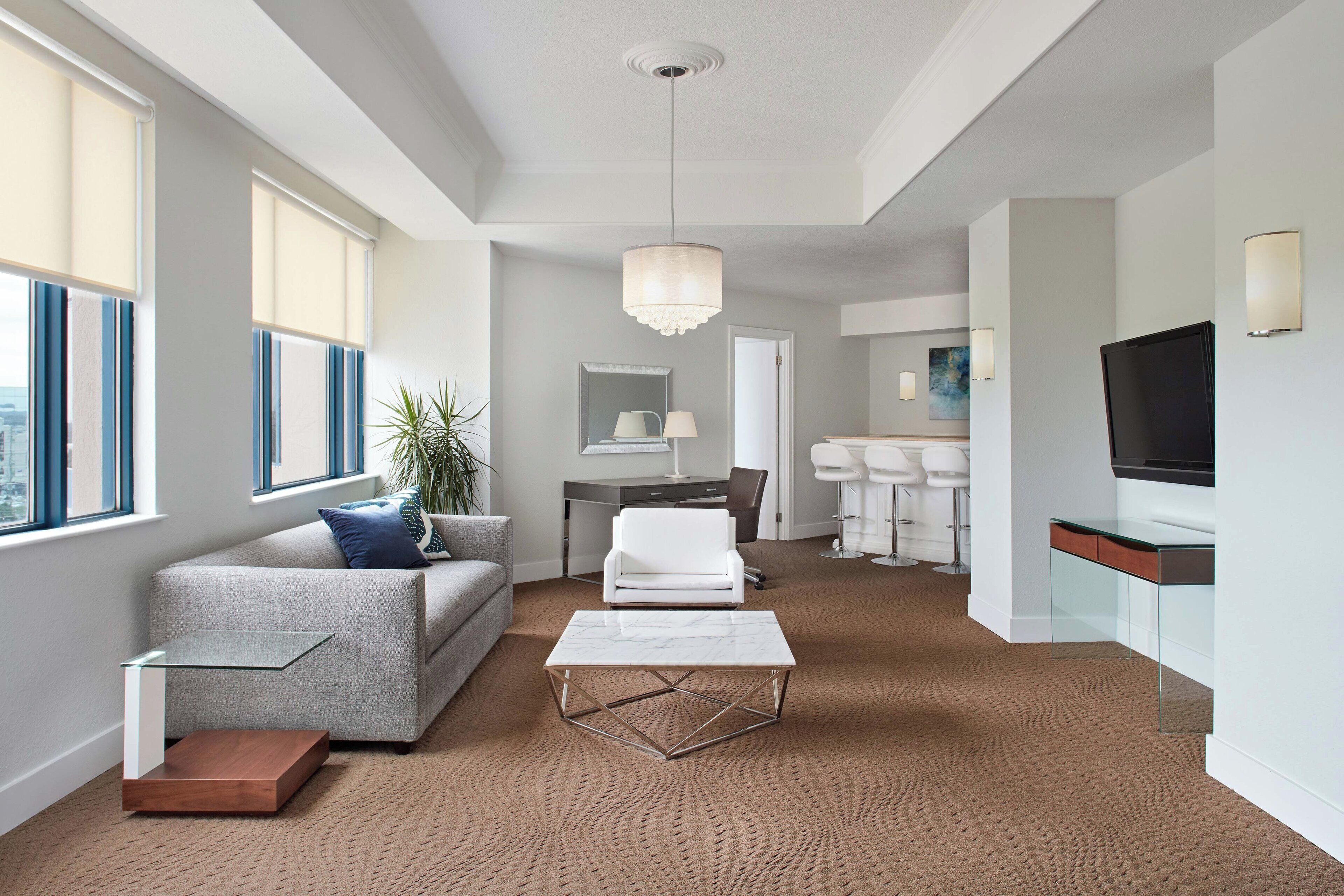 Una habitación de hotel moderna con un sofá gris, un sillón blanco, una mesa de centro de vidrio, un espejo grande y un televisor.