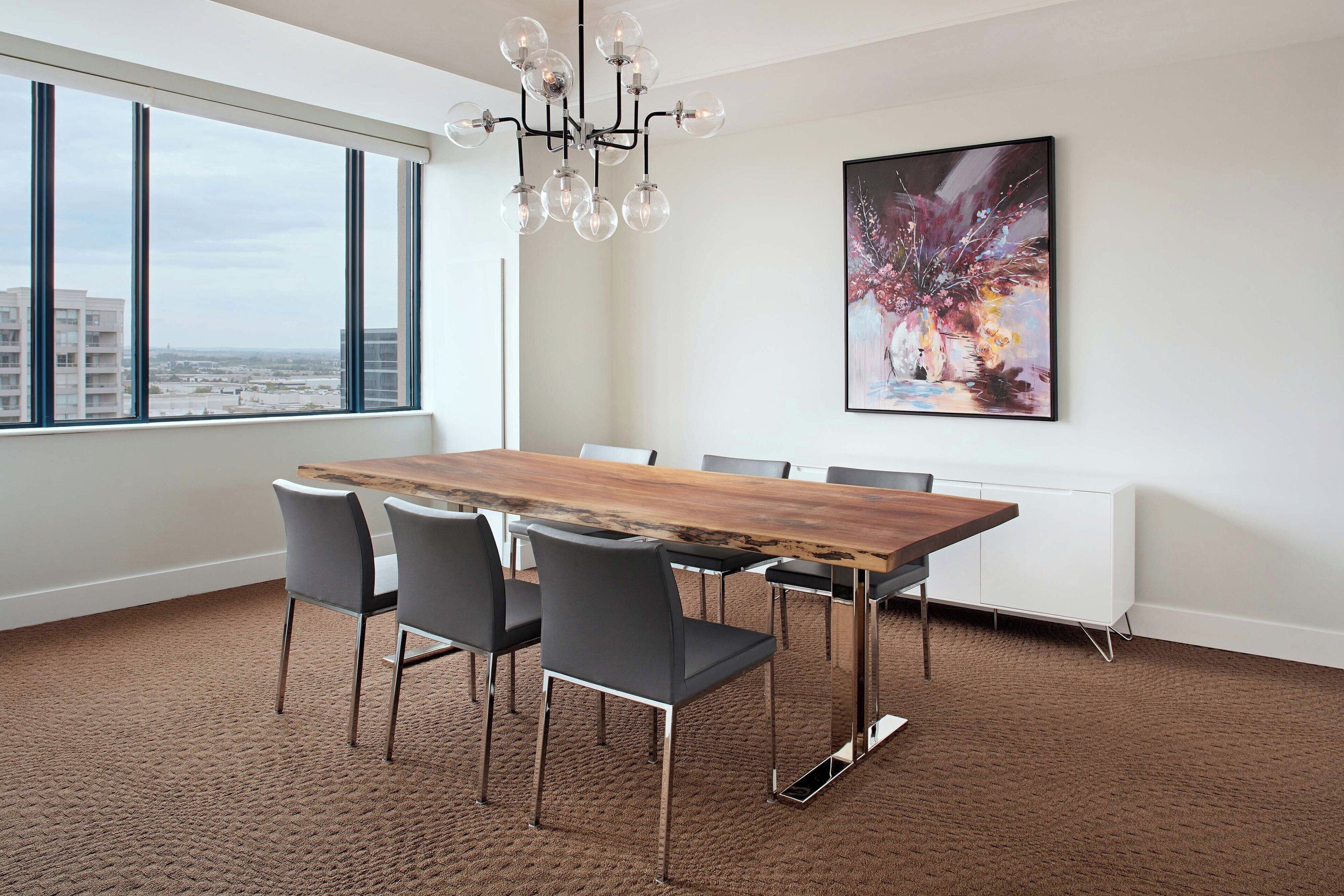 Uma sala de jantar moderna com uma grande mesa de madeira, cadeiras pretas, um candelabro e vista para a cidade através de amplas janelas.