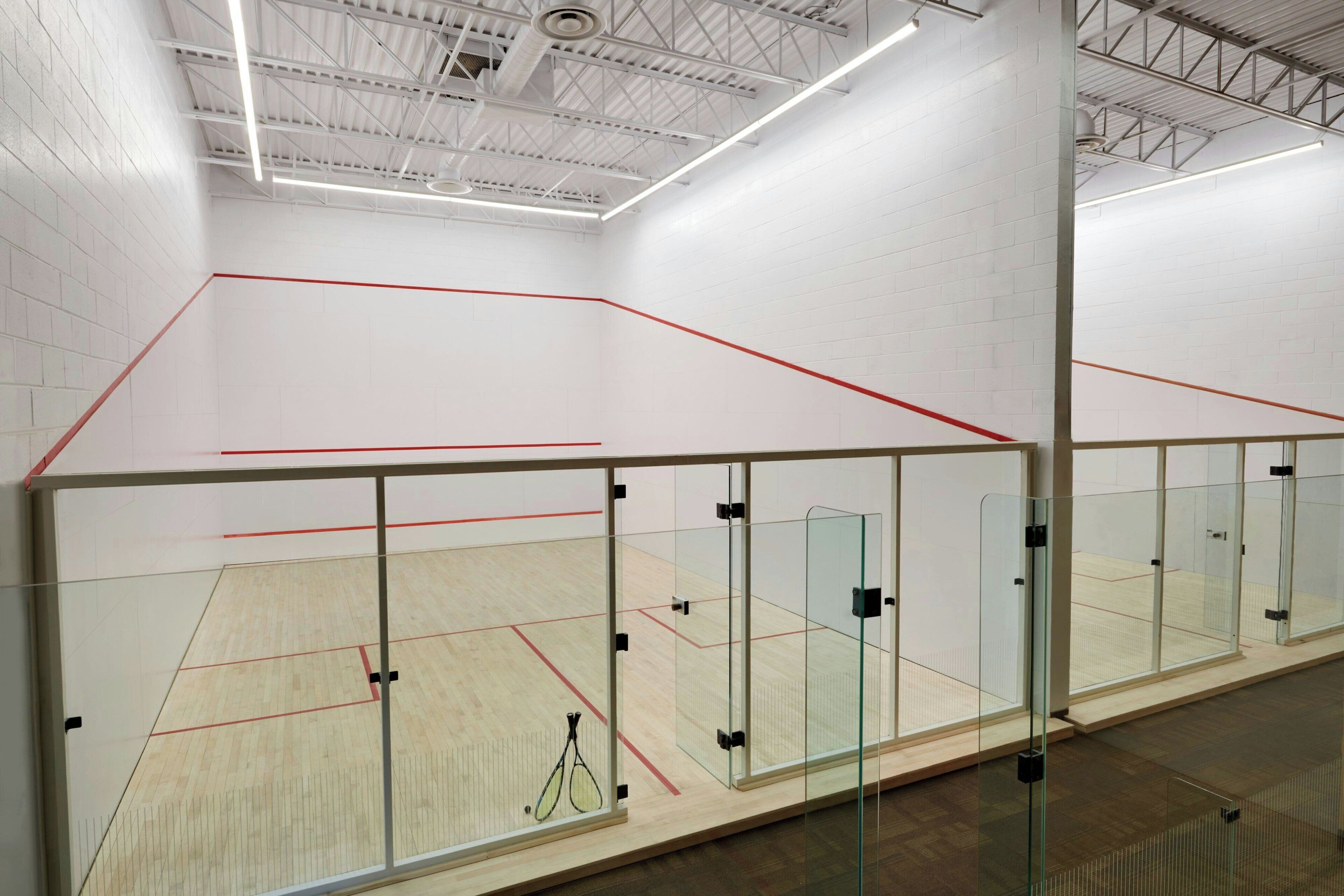A squash court with glass walls and a wooden floor, featuring a red line marking the playing area.
