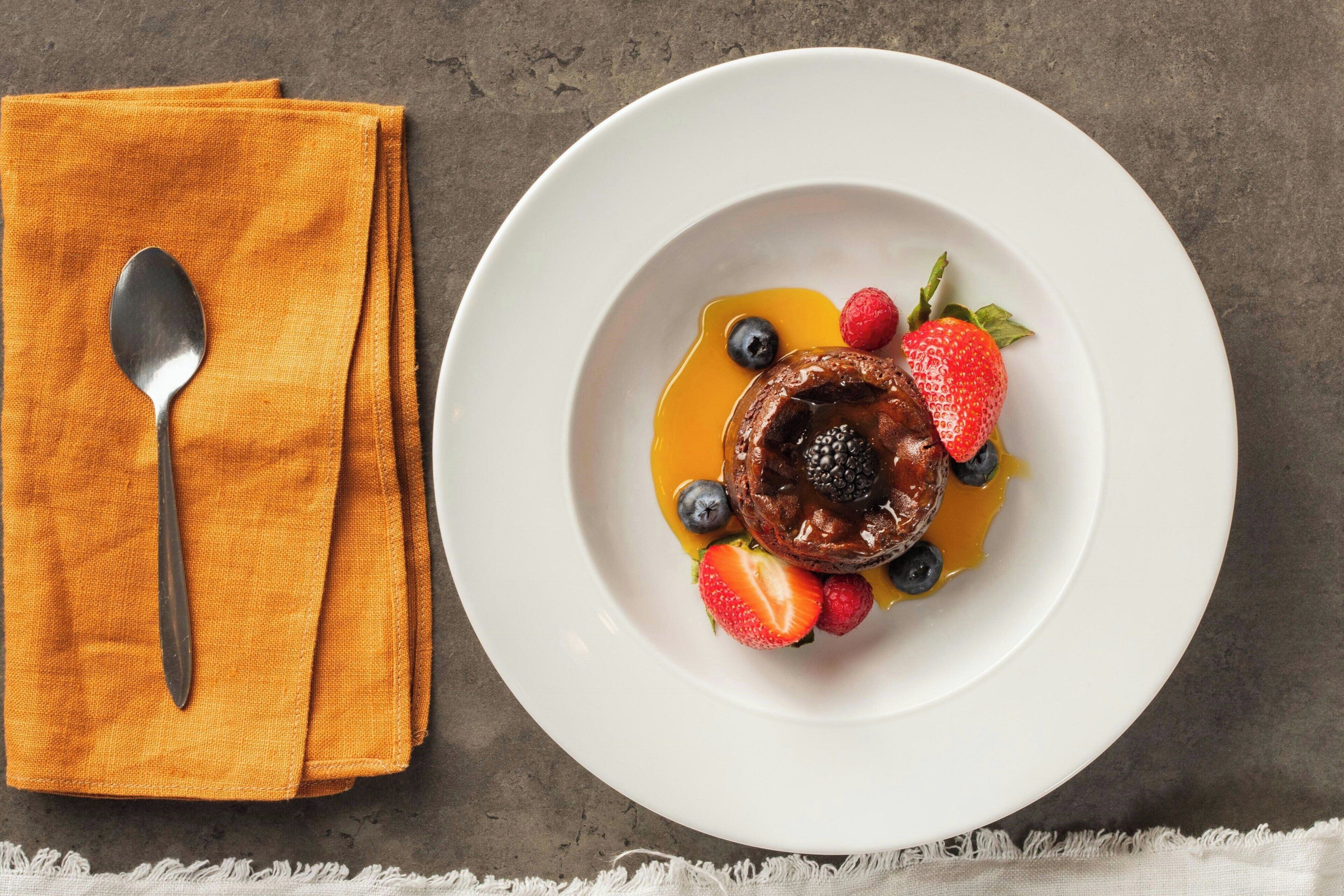 A dessert plate with a chocolate cake, strawberries, blueberries, and a spoon on an orange napkin.