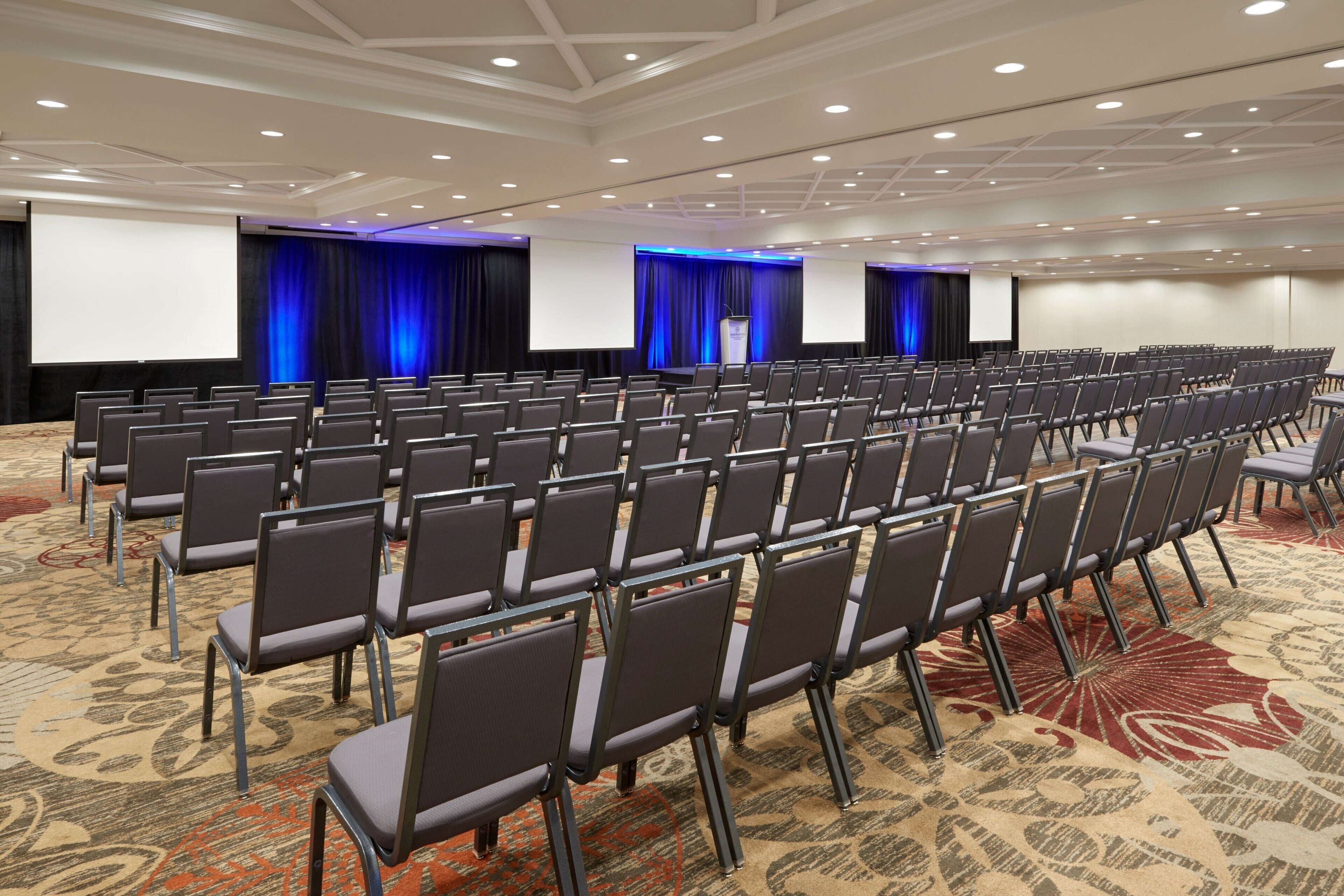 A conference room with rows of chairs, a stage with curtains, and a large screen.