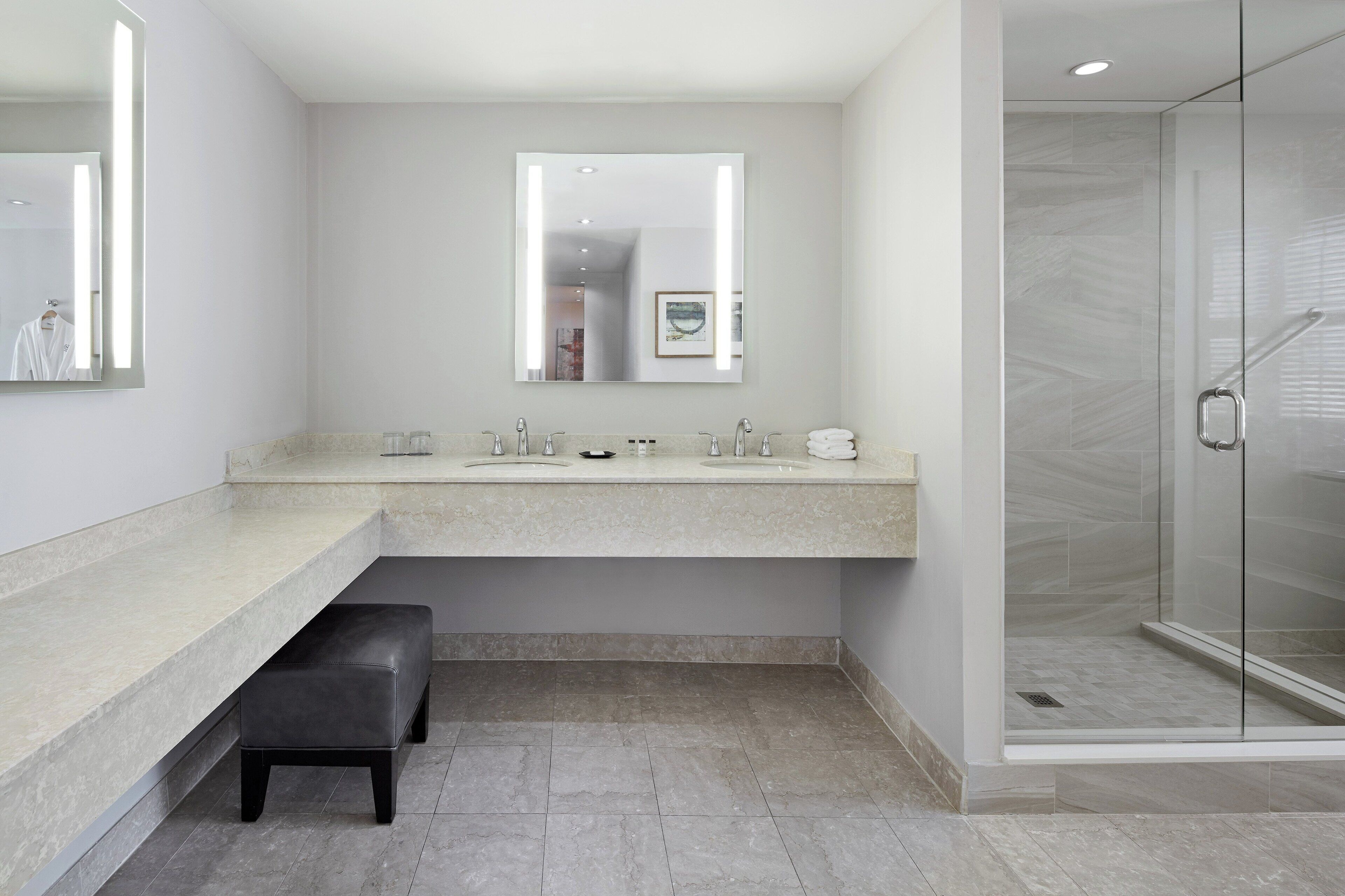 Un baño moderno con un espejo grande, dos lavamanos, un banco y una cabina de ducha de vidrio.