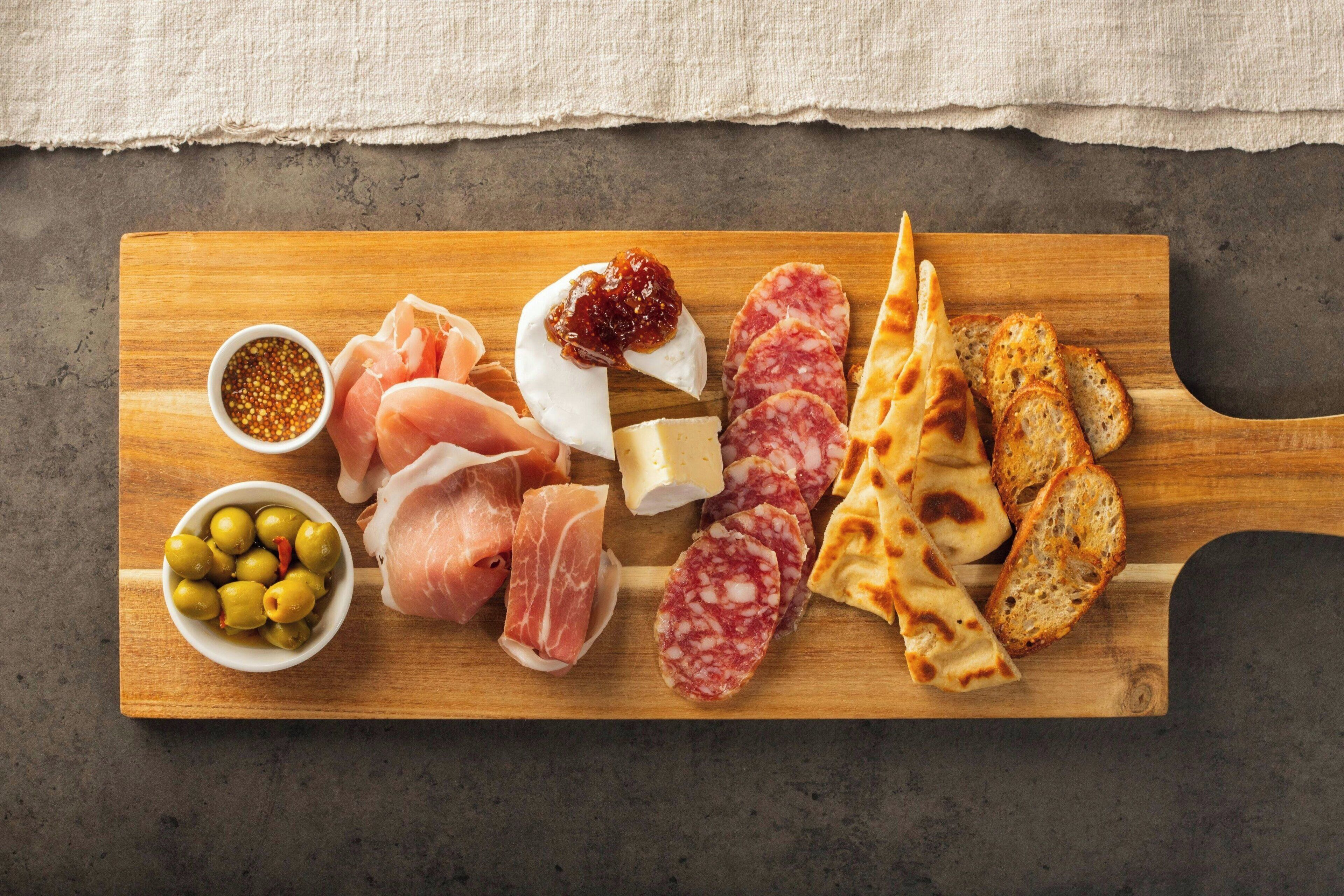 A wooden board with various food items including meats, cheese, olives, and bread.