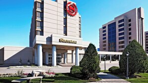 Un hotel Sheraton con un logotipo rojo llamativo, una entrada cubierta y un jardín ajardinado con una banca.
