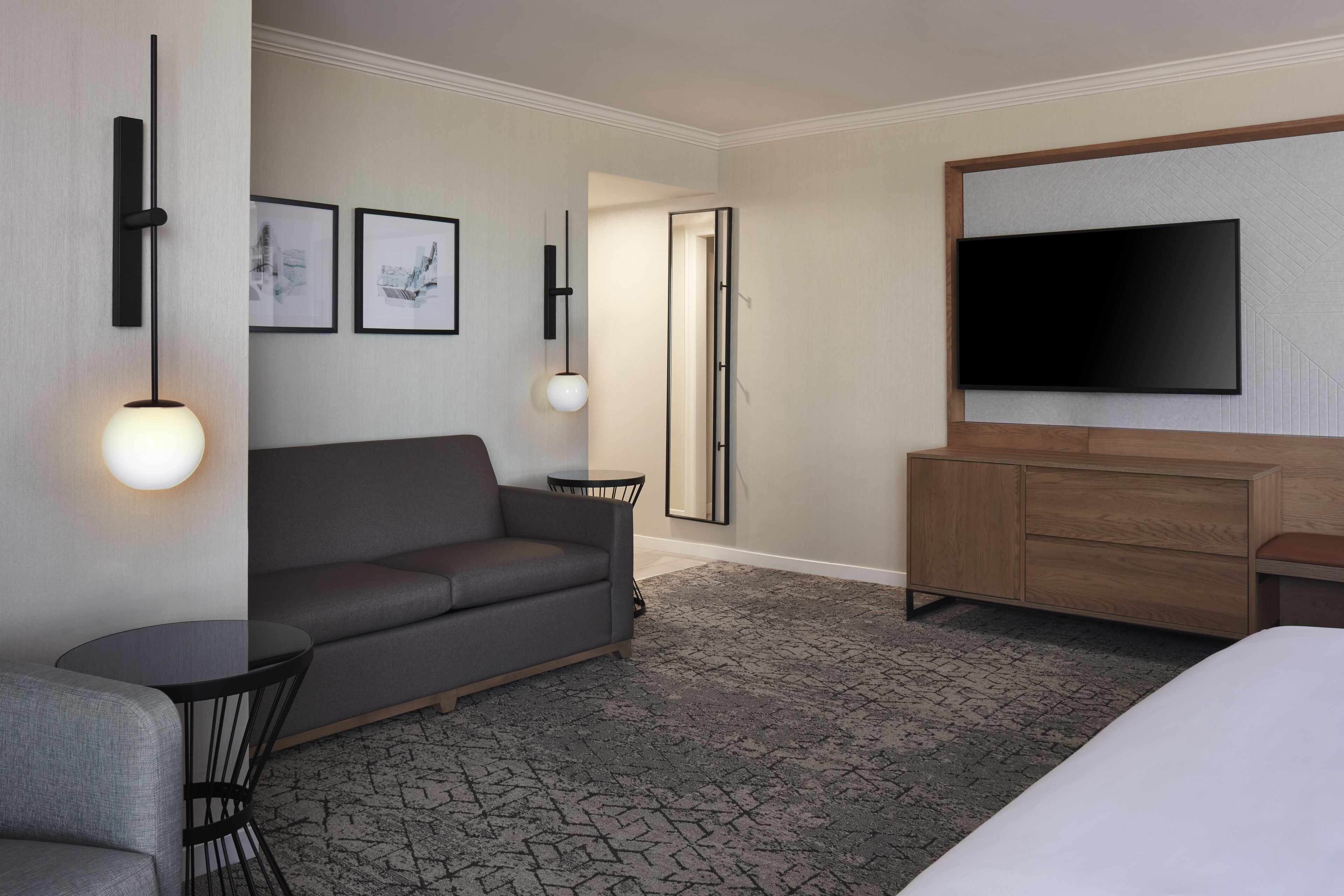 A hotel room with a gray sofa, a small round table, a TV mounted on the wall, and a wooden cabinet.