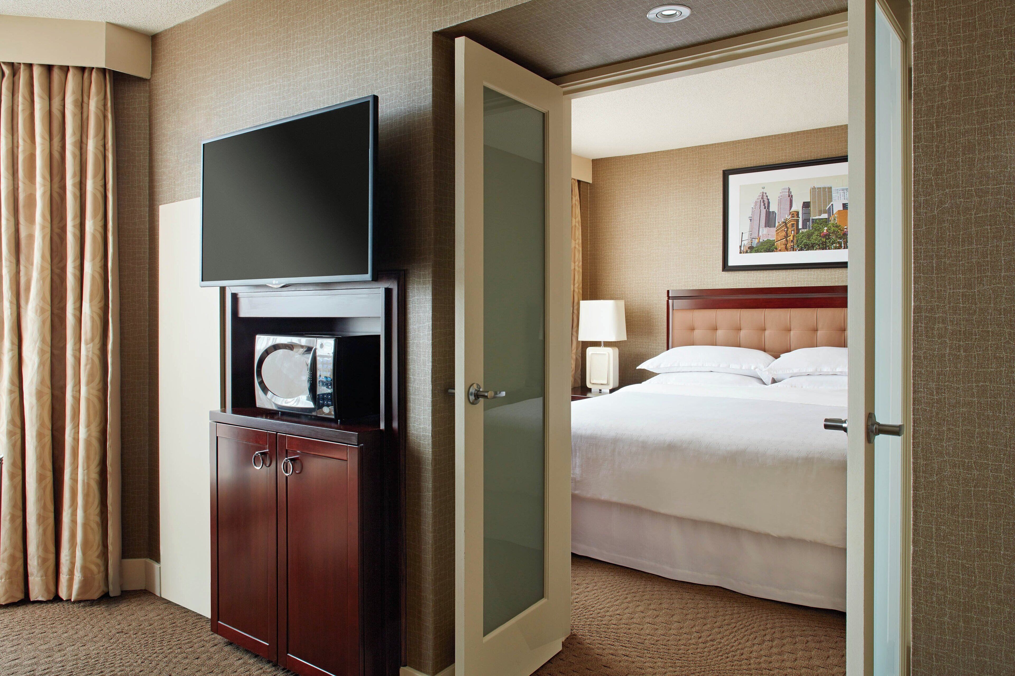 A hotel room with a bed, a television, a minibar, and a cityscape painting on the wall.