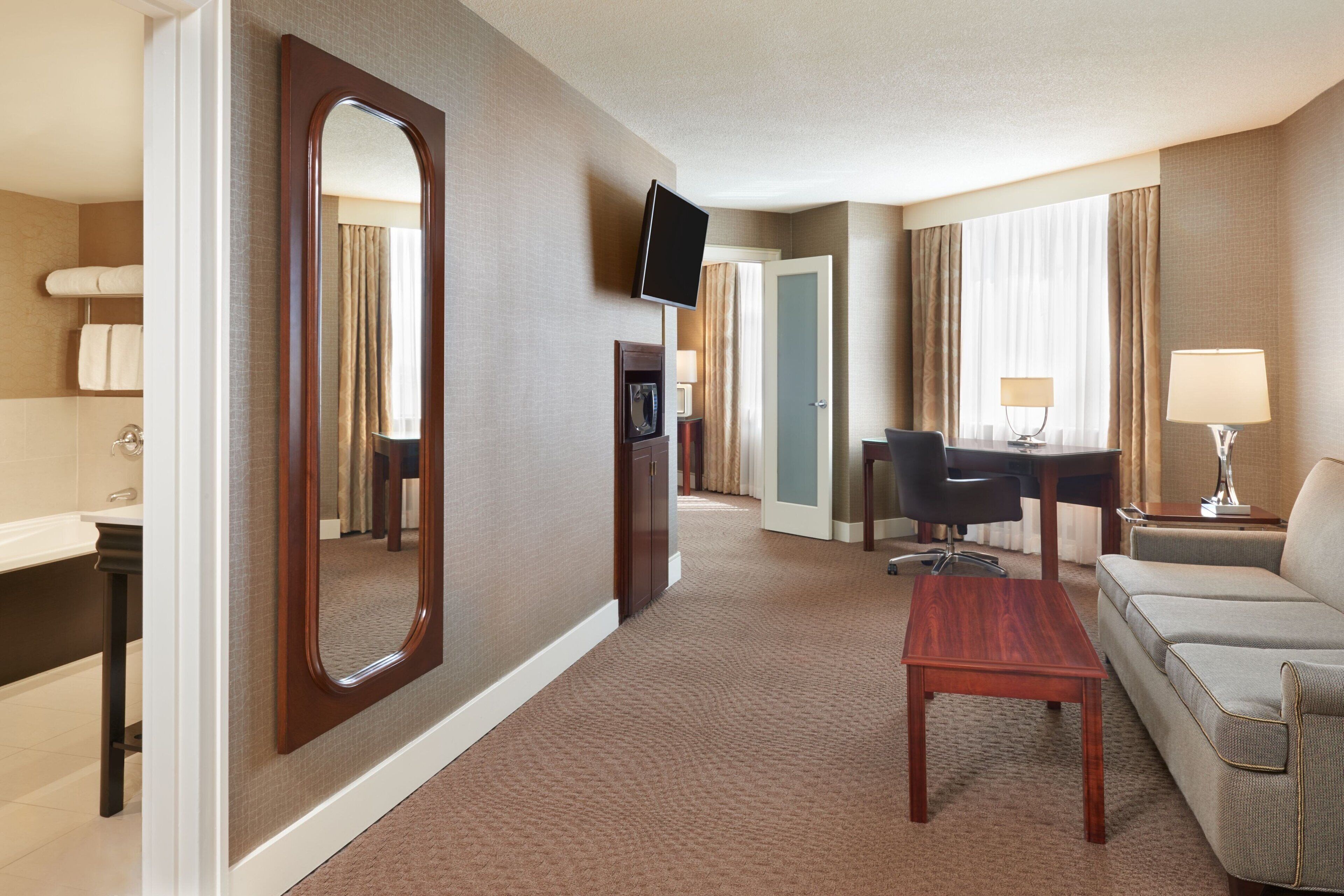 Une chambre d’hôtel dotée d’un grand miroir, d’un téléviseur à écran plat, d’un canapé, d’un bureau et d’une petite table.
