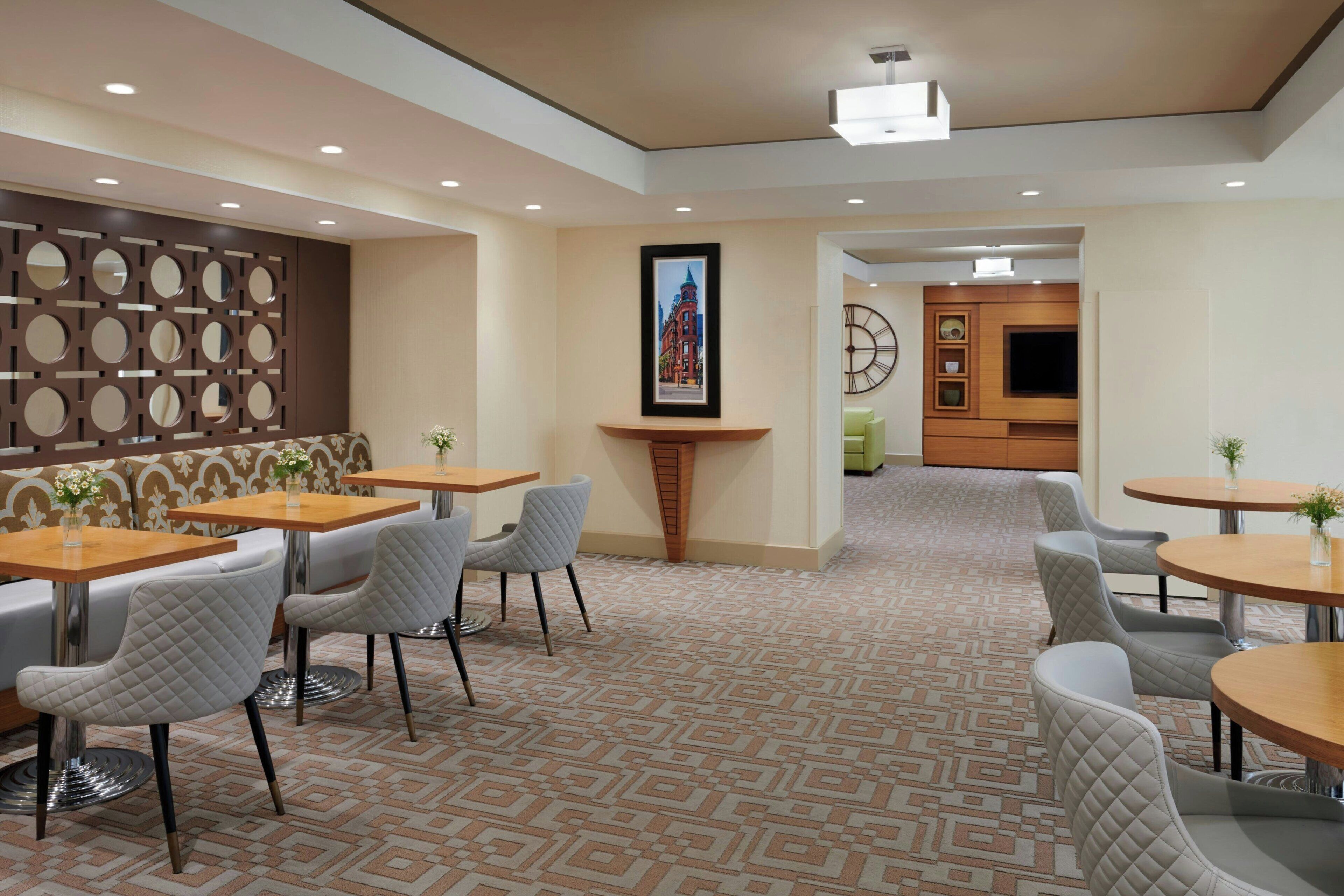 Un vestíbulo de hotel con alfombra estampada, zonas de estar con mesas y sillas, una pared decorativa con espejos circulares y un cuadro enmarcado.