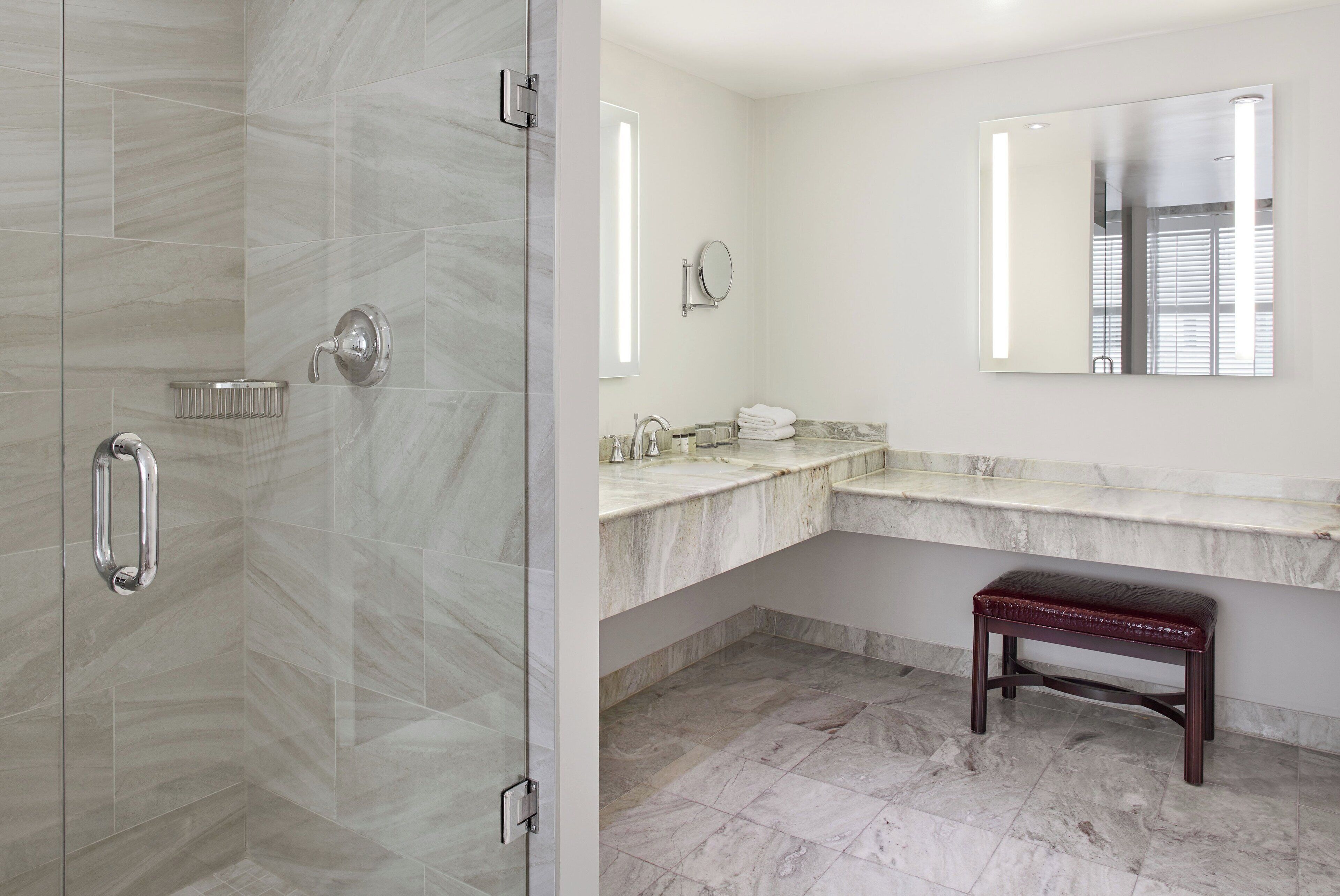 Un baño con cabina de ducha de vidrio, lavabo de mármol, tocador y banco.