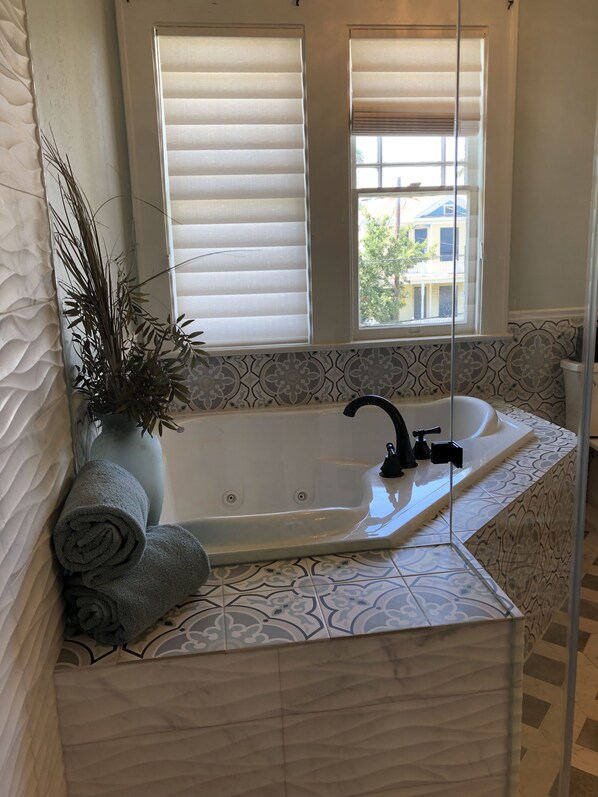 A bathroom with a large bathtub, a window with blinds, a potted plant, and rolled towels. - Avenue O Bed & Breakfast (Galveston)