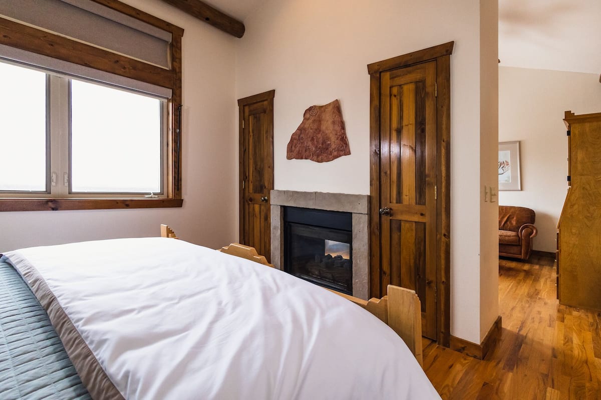 A bedroom with a large bed, a fireplace, wooden doors, and a window.