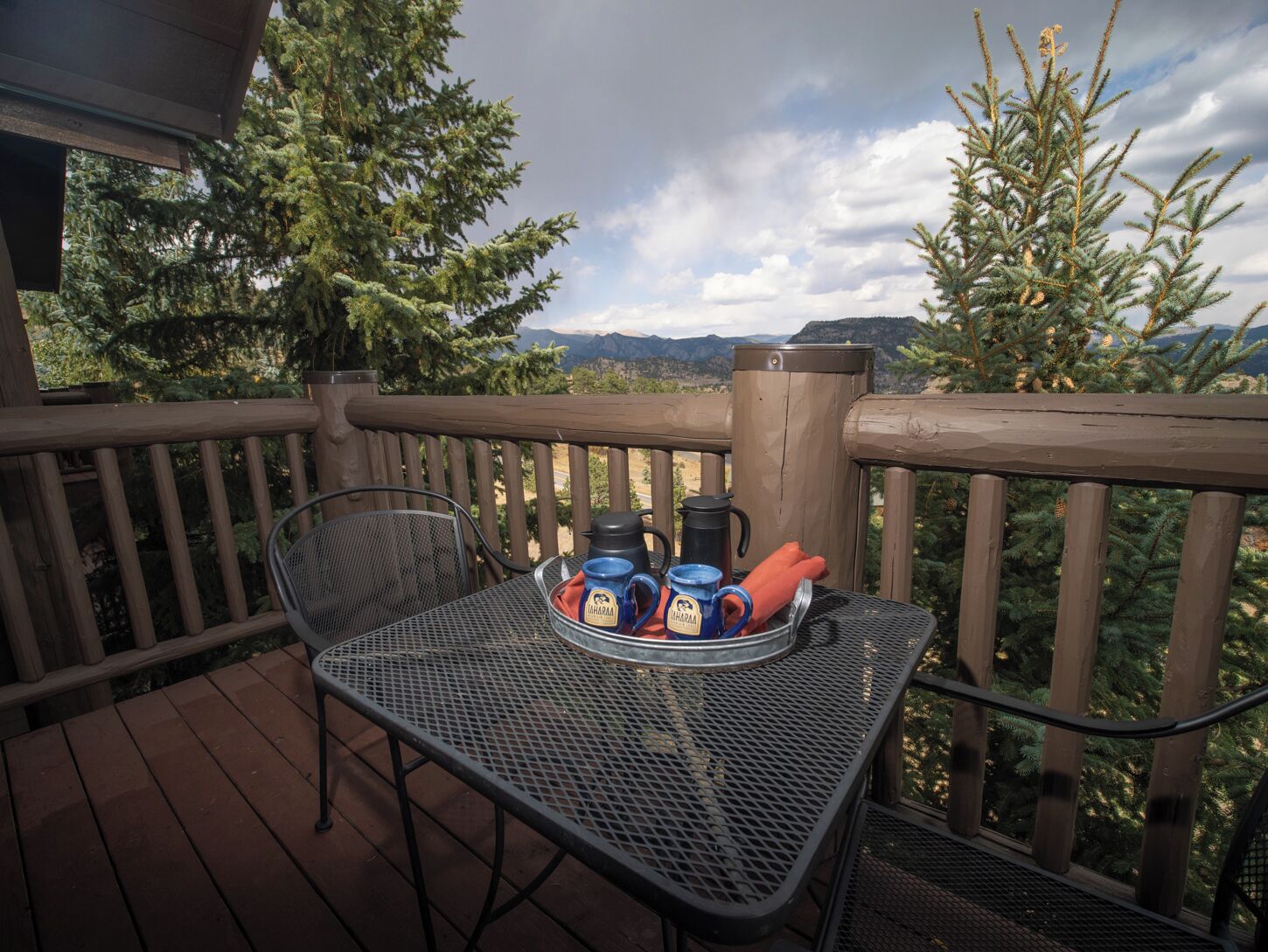 Ein Metalltisch mit einer Tablett voller Tassen und einem Thermos auf einer Holzterrasse mit Geländer, mit Blick auf eine Berglandschaft.