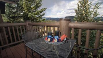 Ein Metalltisch mit einer Tablett voller Tassen und einem Thermos auf einer Holzterrasse mit Geländer, mit Blick auf eine Berglandschaft.