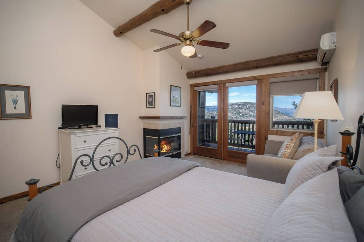 A bedroom with a bed, a television on a dresser, a fireplace, and a balcony with a view of snow-covered hills.
