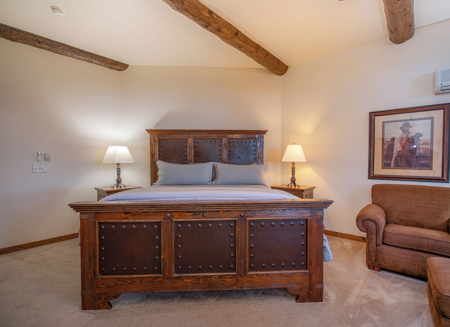 Een slaapkamer met een groot houten bed, twee bedlampen, een ingelijst schilderij aan de muur en een bruine fauteuil.