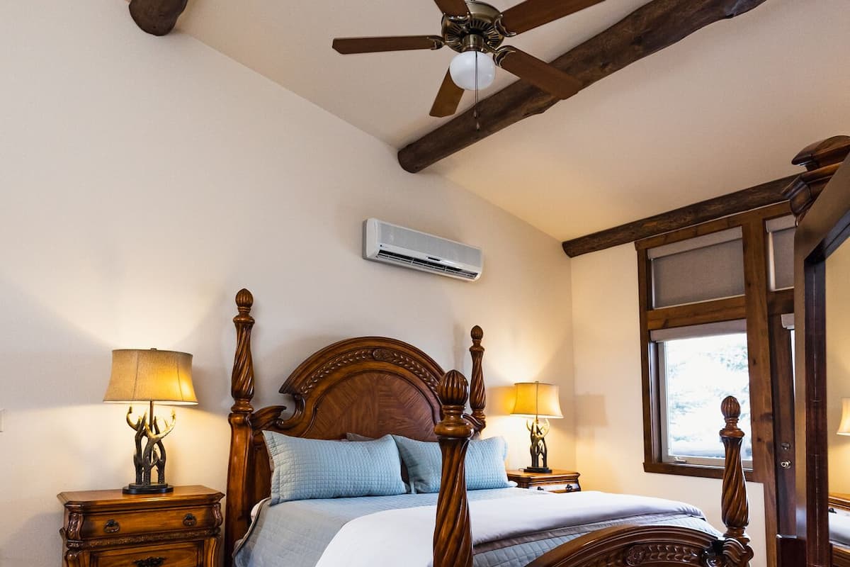 A bedroom with a wooden four-poster bed, two bedside tables with lamps, a ceiling fan, and a window with a view.