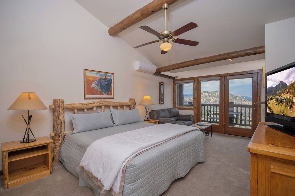 A bedroom with a bed, two bedside tables, a ceiling fan, a TV, and a balcony with a mountain view.