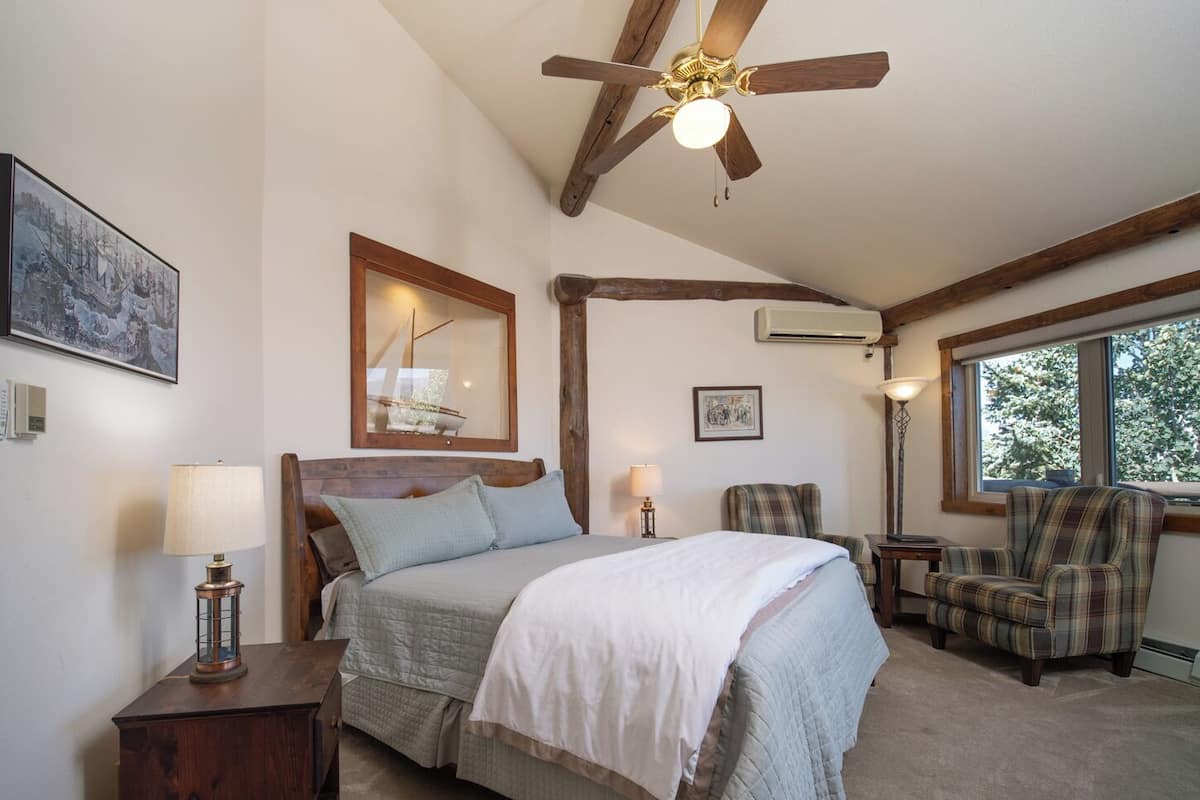 A bedroom with a bed, two chairs, a nightstand, a ceiling fan, and a window.