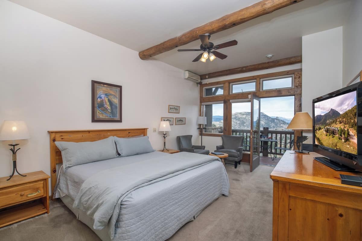 A bedroom with a large bed, a ceiling fan, a TV, and a balcony with a mountain view.