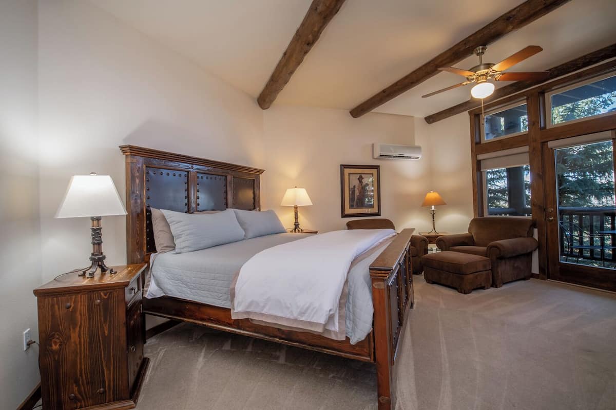 A bedroom with a wooden bed, two bedside tables with lamps, a chair, a ceiling fan, and a large window with a view of trees.