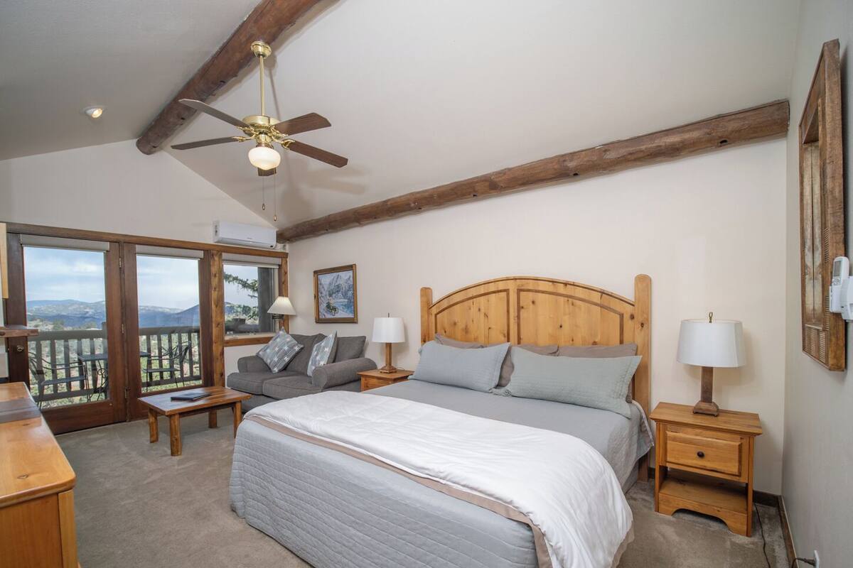 A bedroom with a large bed, a wooden headboard, two bedside tables with lamps, a nightstand, a window with a view of mountains, and a ceiling fan.
