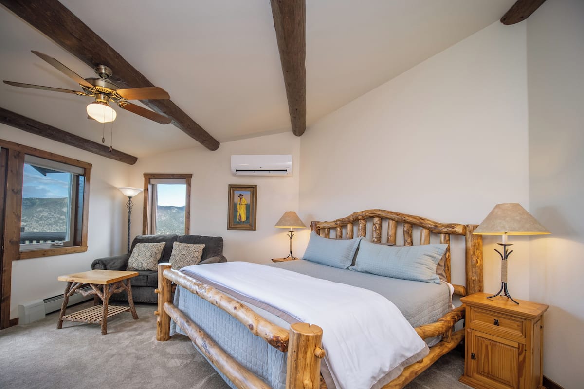 A bedroom with a wooden bed frame, two bedside tables with lamps, a small wooden table, a sofa, and a window with a view.