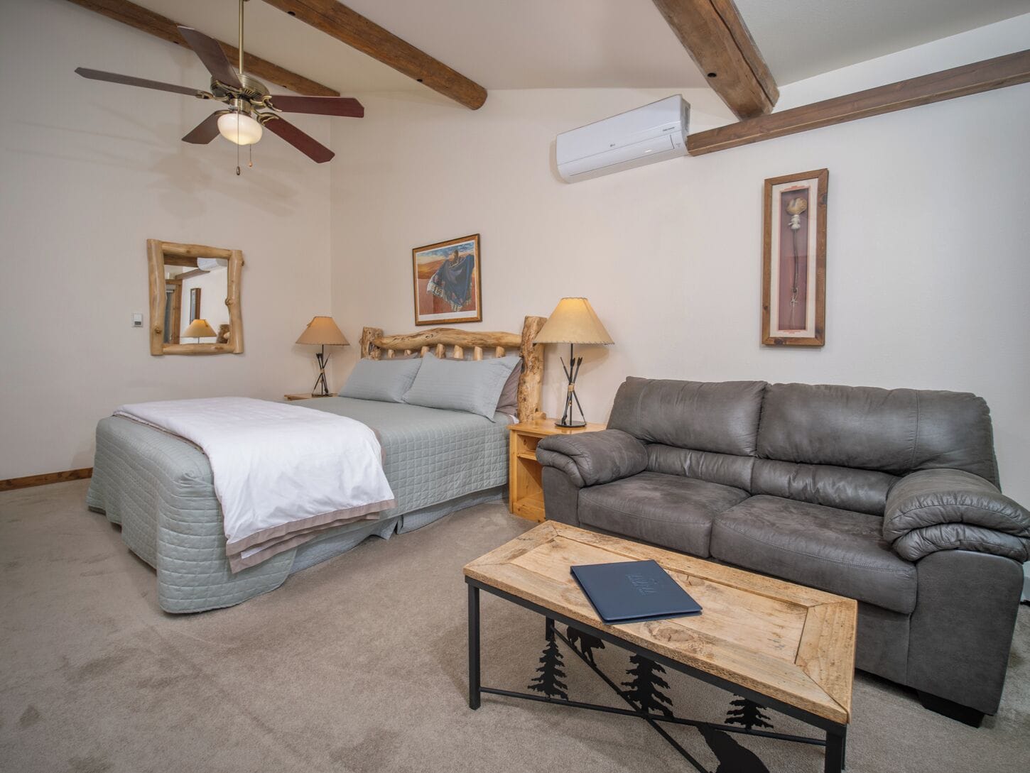 A bedroom with a bed, a gray leather sofa, a wooden coffee table, and a ceiling fan.
