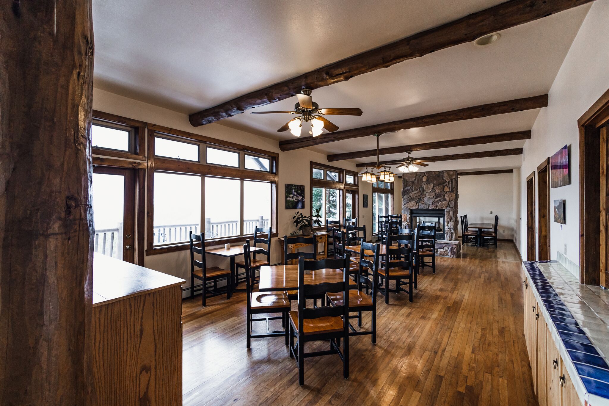 Une salle à manger spacieuse, avec des tables et des chaises en bois, de grandes fenêtres et une cheminée en pierre.