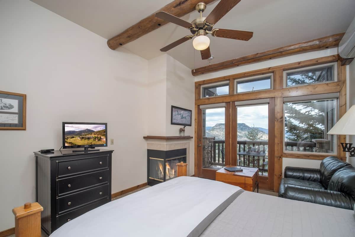 A bedroom with a bed, a dresser, a television, a fireplace, and a balcony with a view of mountains.