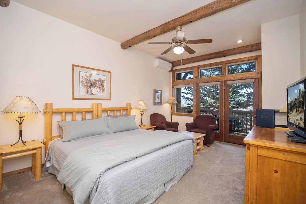 A bedroom with a bed, wooden headboard, two bedside tables with lamps, a chair, a desk with a computer, and a large window with a view of trees.