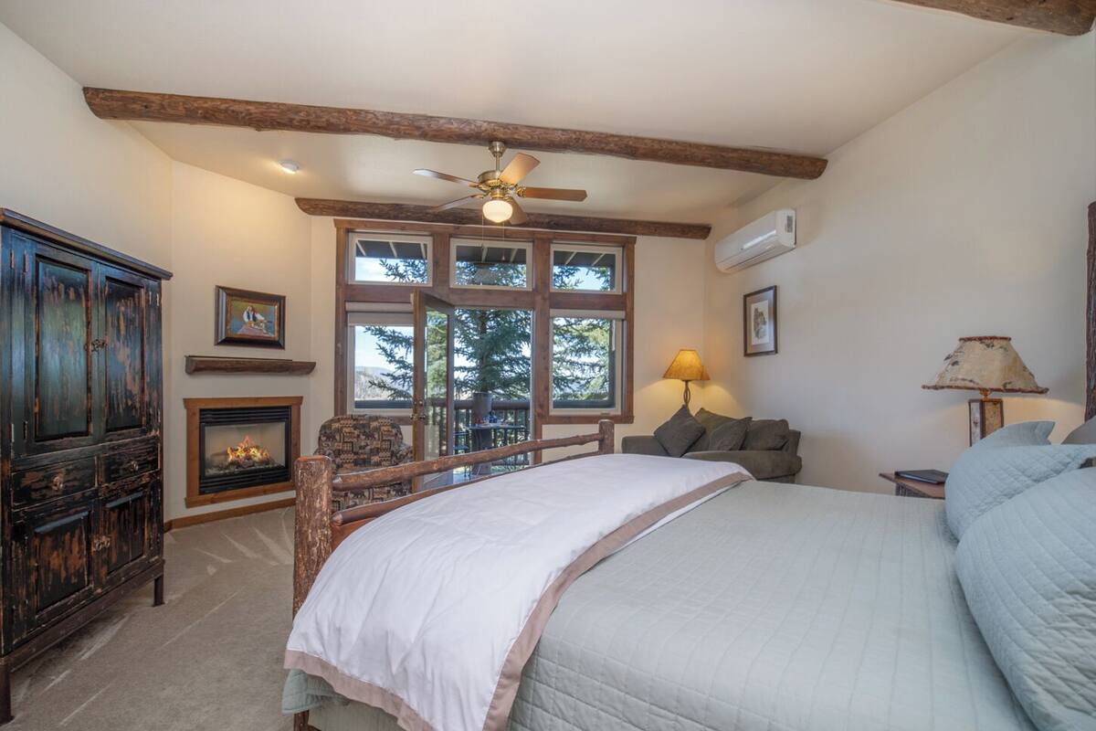 A bedroom with a large bed, wooden beams, a ceiling fan, a fireplace, and a view of trees through the windows.