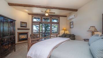 A bedroom with a large bed, wooden beams, a ceiling fan, a fireplace, and a view of trees through the windows.