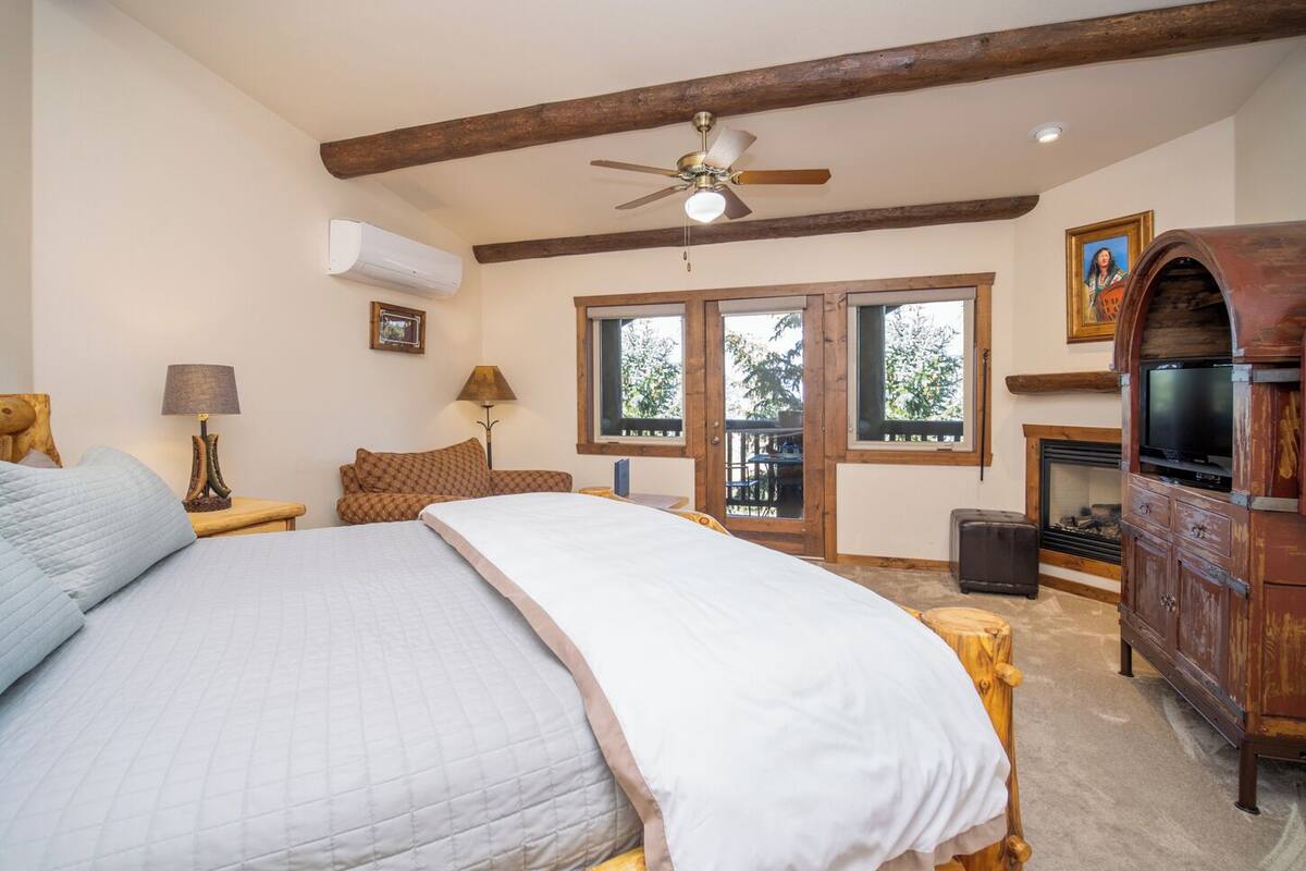 A bedroom with a large bed, wooden beams, a ceiling fan, a fireplace, and a television.