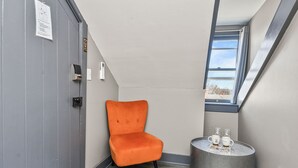 Una habitación en esquina con una puerta gris, una silla pequeña color naranja, una mesa redonda con dos vasos y una ventana con vistas.