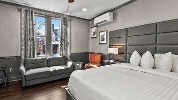 A hotel room with a bed, a grey sofa, a chair, a bedside table with a lamp, and a window with curtains.