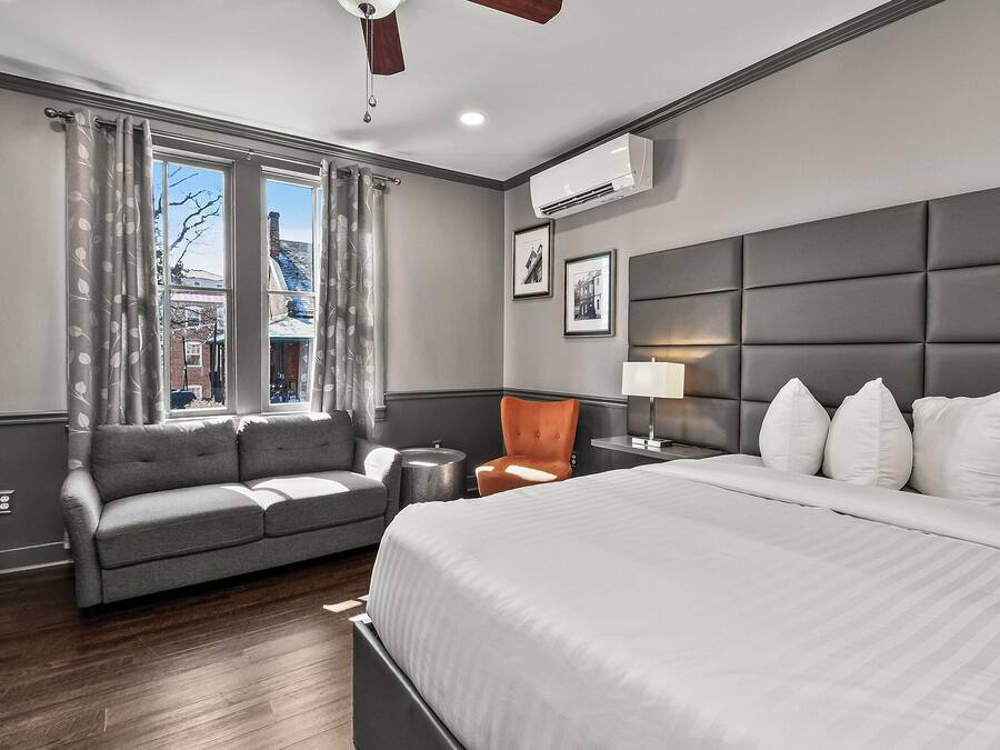 A hotel room with a bed, a grey sofa, a chair, a bedside table with a lamp, and a window with curtains.