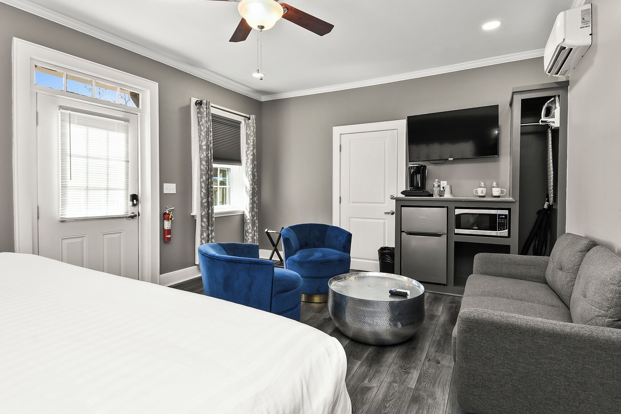 A hotel room with a bed, two blue armchairs, a gray sofa, a round coffee table, a kitchenette with a microwave, and a ceiling fan.