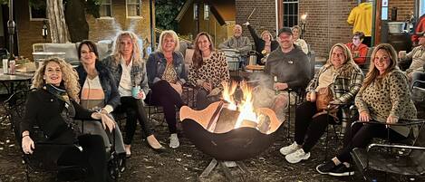 A group of people gathered around a fire pit in an outdoor setting with string lights overhead.