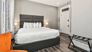 A modern hotel room with a large bed, a nightstand with a lamp, an orange chair, and a metal rack.
