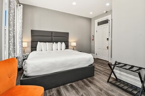 Una habitación de hotel moderna con una cama grande, una mesita de noche con lámpara, un sillón naranja y un perchero metálico.