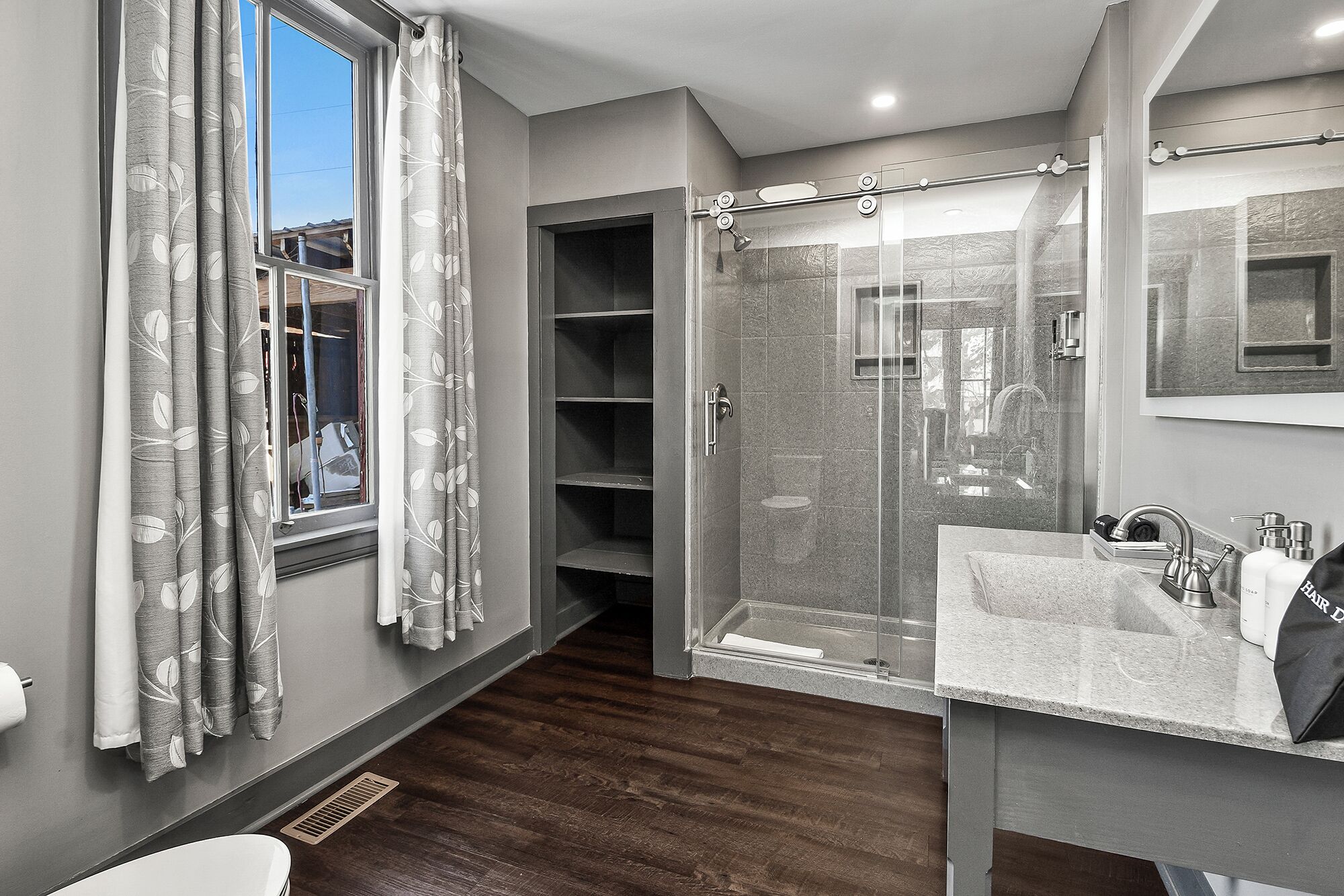 Un baño moderno con ducha de vidrio, tocador con lavamanos y espejo, y ventana con cortinas.