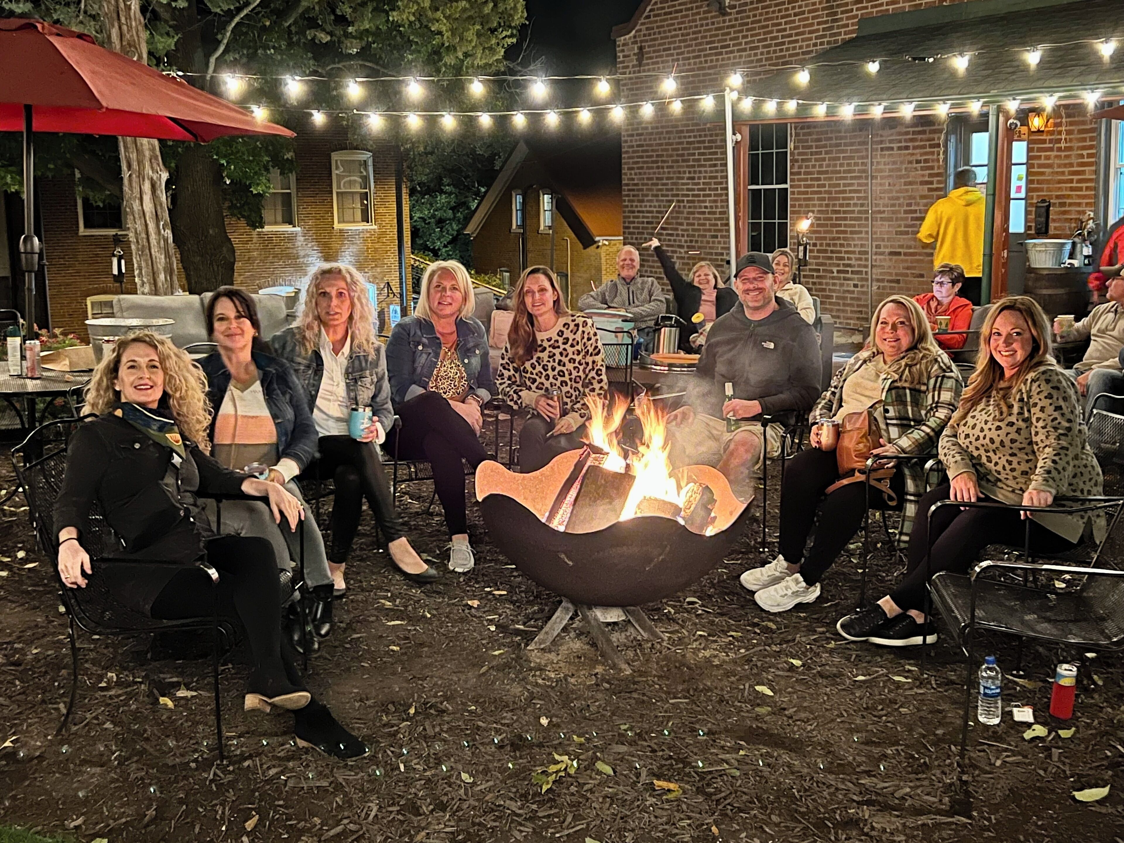 Un grupo de personas reunidas alrededor de una fogata en un entorno al aire libre, con luces colgantes sobre ellas.